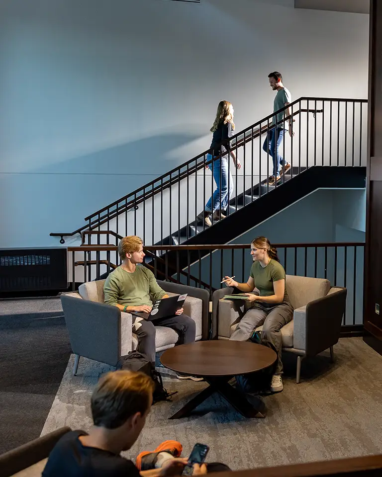 three students sit in lounge while two students walk on stairs