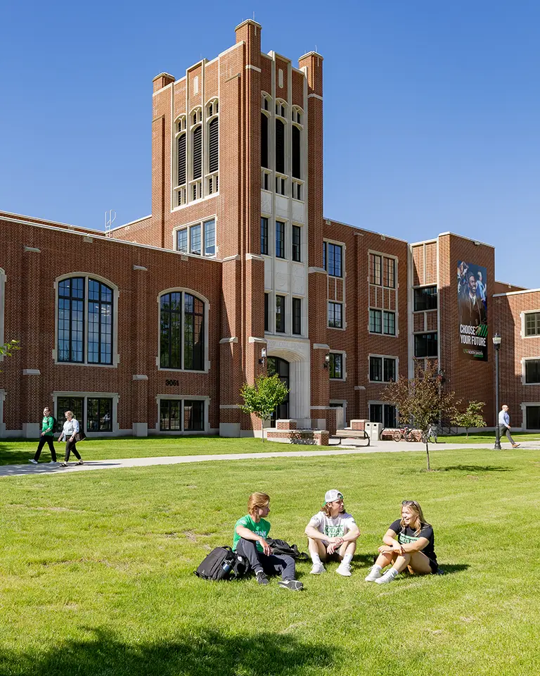 Three students sitting in grass in front of library building