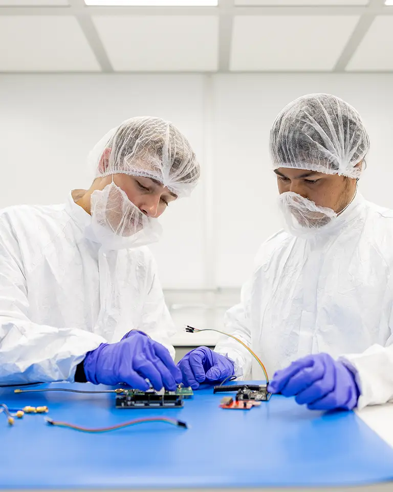 Two students wearing all white and hair nets connect electrical panels in a clean room.