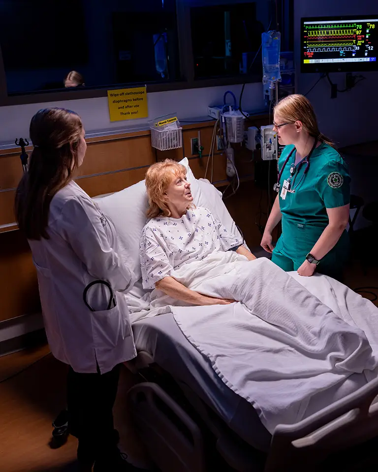 A med student and a nursing student talk to a person in a hospital bed simulator.