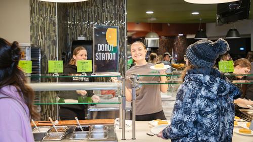 Student dining worker smiles at the camera as she hands a student a plate of food from behind the service counter