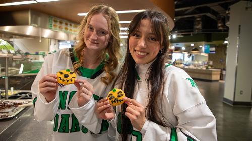 Two female students in white North Dakota hockey jerseys pose with frosted sugar cookies decorated to look like Halloween jack-o-lanterns