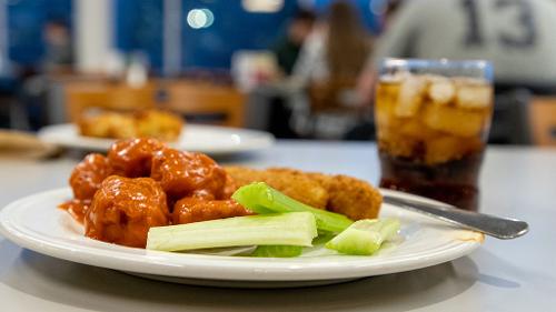 Plate of classic buffalo wings with celery sticks on a table with a glass of iced cola; dining center and patrons blurred in the background