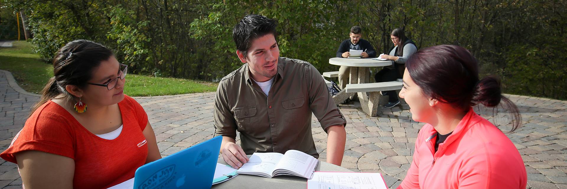 Three students are seated at a table engaged in conversation. They have have a book, notepad, and computer in front of them.