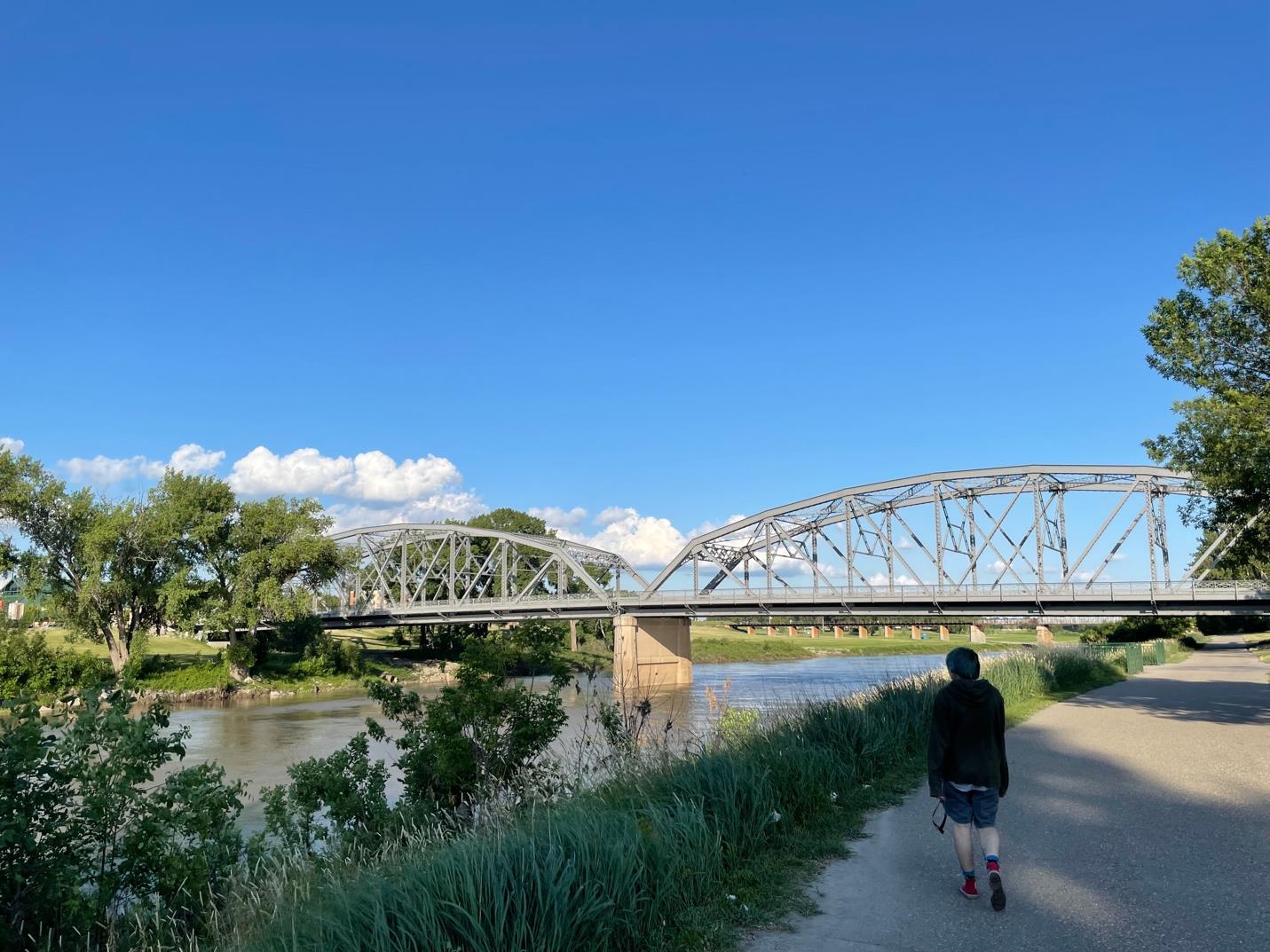 One of the bridges over the Red River in Grand Forks, ND. Behind the bridge is a brilliant blue sky with a few clouds hovering nearer the horizon. In the foreground, a person is walking along a path toward the bridge. 