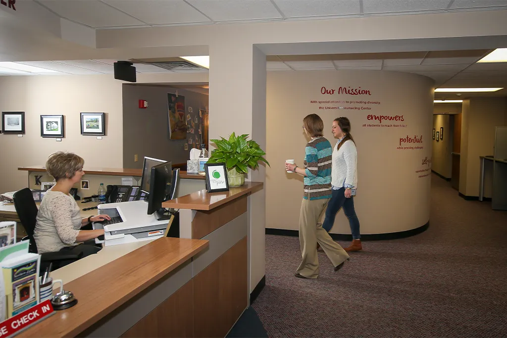 Two students walk past the University Counseling Center reception desk and mission statement wall.