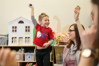 A special education teacher interacts with a young boy in a classroom. The boy raises his hand excitedly while holding a blue ball, and the teacher holds a similar object