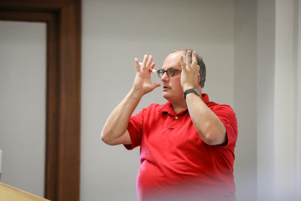 In a lecture hall, a psychology professor stands with enthusiasm, gesturing emphatically with both hands near his face as he delivers an engaging lecture