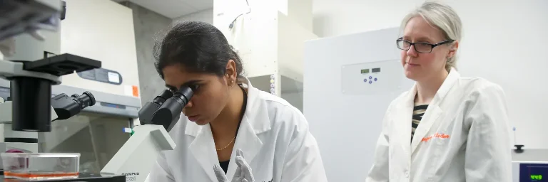 Biomedical engineers in a lab: one peers through a microscope while the other observes closely, surrounded by scientific equipment