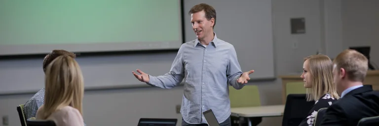 A marketing professor giving a lecture to their students in a classroom, gesturing with hands to emphasize key points