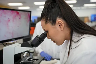 A medical laboratory science student looks into a microscope, the magnified sample shown on a monitor.