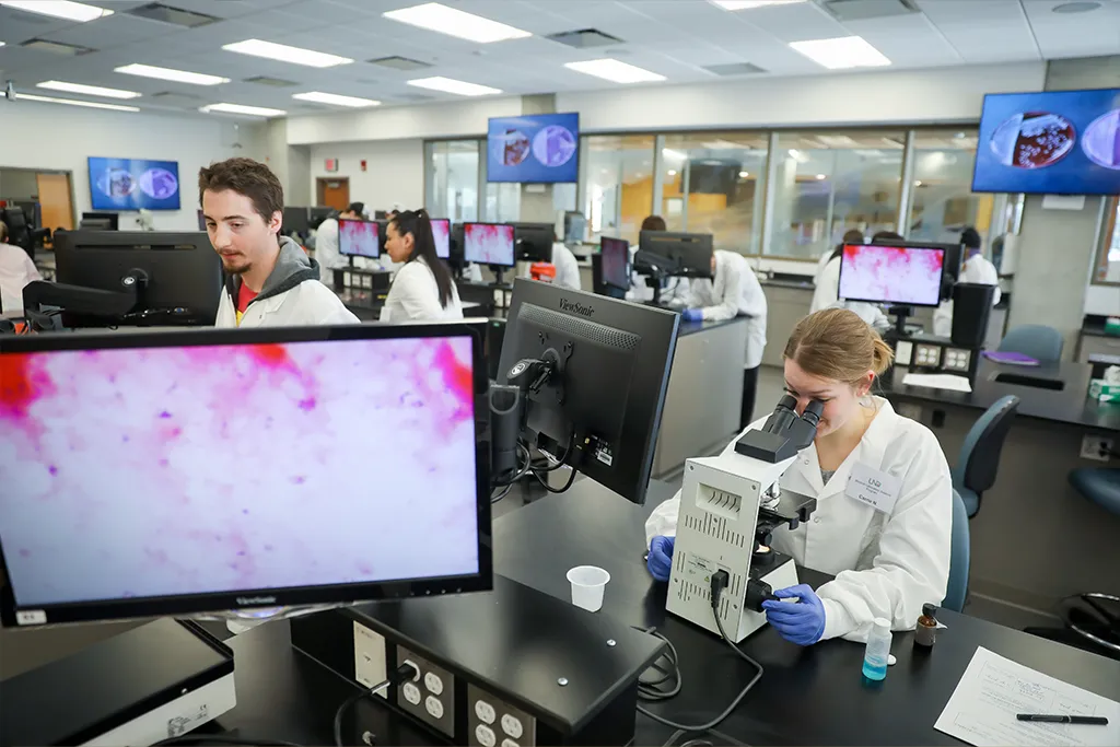 Medical laboratory science students examine samples at microscope stations with cells shown on monitors.