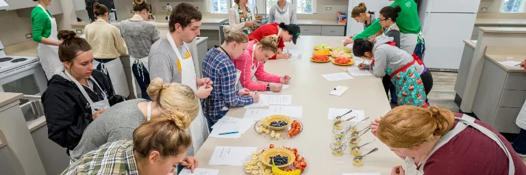 Nutrition students in a cooking class, preparing dishes with ingredients like bananas, blueberries, and yogurt, while also writing down the calorie count of their creations