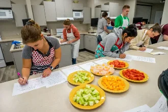 Students in a nutrition class gather around a table, pens in hand, as they study the calories of the fruits laid out before them