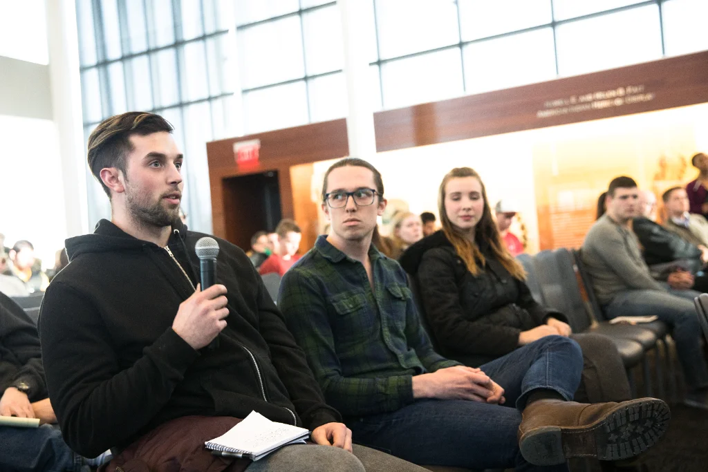 A political science student is speaking into a microphone in a lecture hall, while fellow students listen attentively.