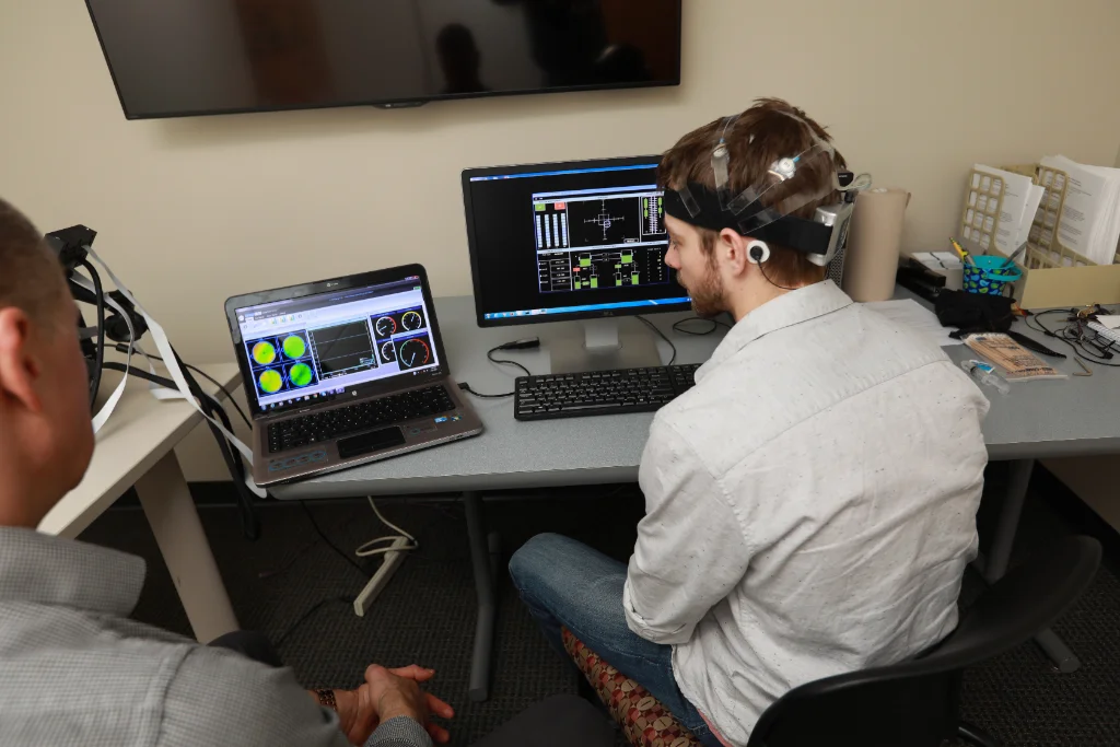 On a computer, a psychology researcher and a student examine brain activity data, while the student wears a sensor cap for monitoring