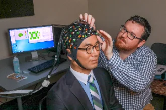 A psychology researcher adjusts a multi-colored sensor cap on a student's head, used for brain monitoring. In the background, a computer screen displays data and graphical representations of brain activity