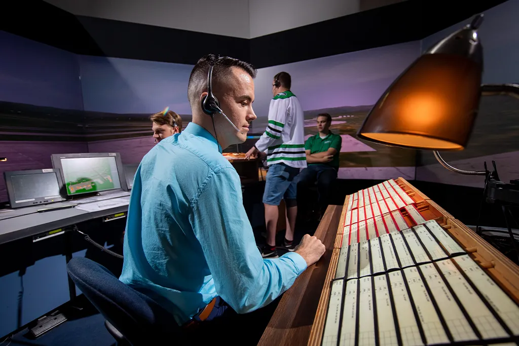An air traffic control student in a headset works flight progress strips at a tower simulator station.
