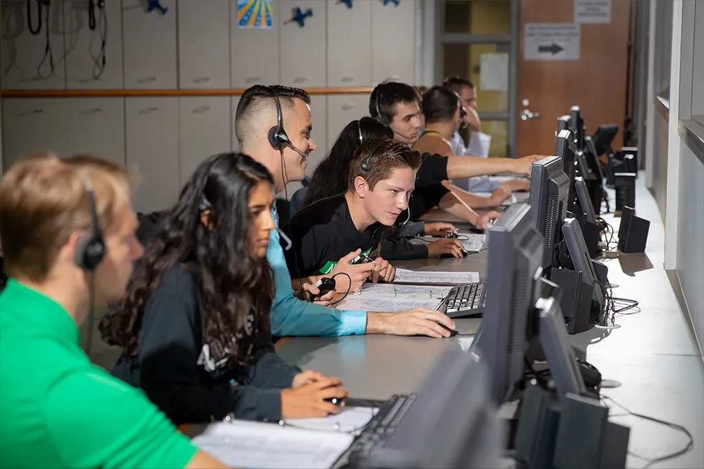 A row of air traffic control students wearing headsets work at computer stations along a long desk.