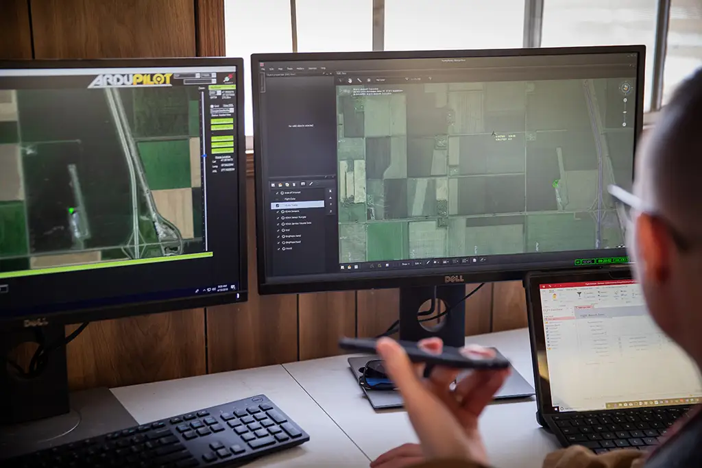 Person monitoring satellite imagery and location data on dual computer screens while holding a phone, representing AI-powered surveillance and the ethical concerns around privacy and tracking