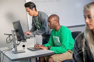 Two students work at desktop computers in a classroom, while another student sits in the foreground, slightly out of focus.