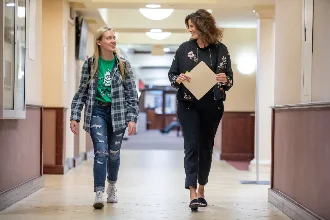 A communication student walks down a hallway with her professor, both engaged in a friendly conversation