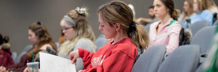 Female psychology student in a red sweatshirt sitting in class, looking at her notes while other students focus on their own materials