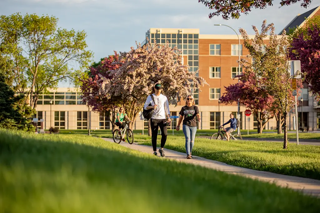 Students walking across the University of North Dakota campus with blooming trees and modern academic buildings in the background, symbolizing a supportive learning environment at an accredited university