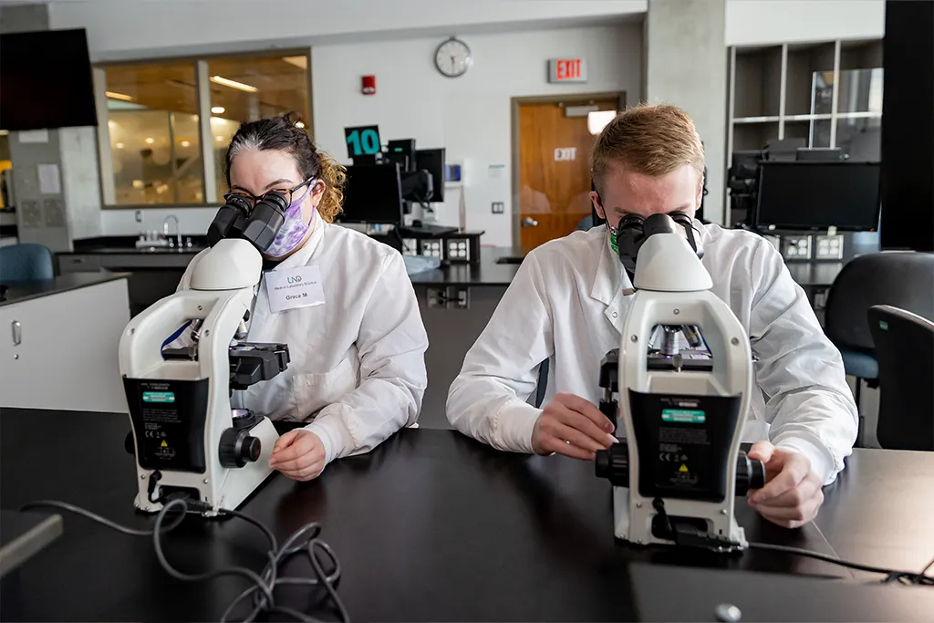 Two medical laboratory science students in white coats and masks examine slides at microscopes.