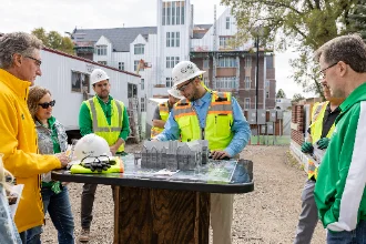 At a construction site, six structural engineers gather around a table with a small architectural model of a building, engaged in collaborative project discussions