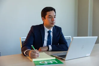 An accountant in a business suit is seated at a desk, writing on a notepad and looking at a laptop screen.