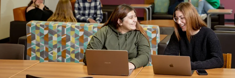 two female cybersecurity students collaborating on a project, using their laptops