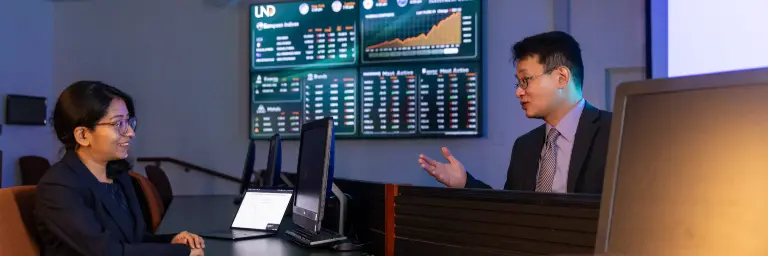 A professor and a student sit together at a desk, analyzing data on a computer screen related to applied economics.