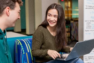 Two students are sitting with their laptops open, laughing