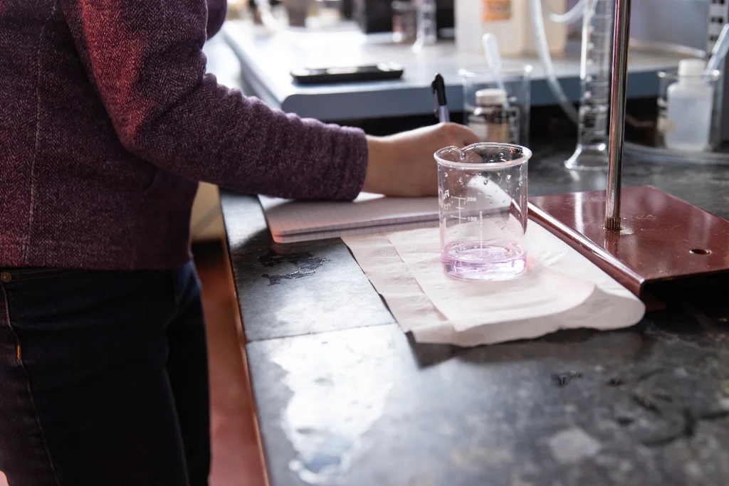 In a chemical engineering laboratory, a student conducts an experiment with a pinkish-purple liquid in a beaker on a black countertop, surrounded by various lab equipment