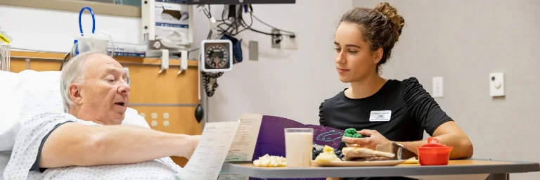 A nutrition degree student observing an elderly man with his meal in a hospital setting, promoting healthy eating habits.