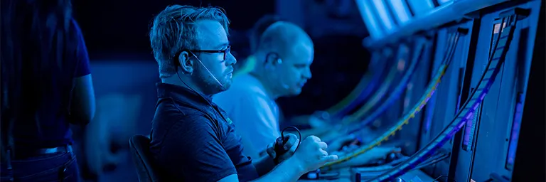 Air traffic control students in headsets monitor radar screens at workstations under blue light.
