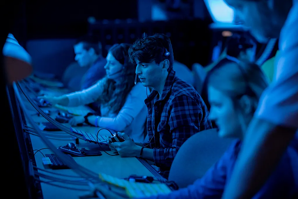 A row of air traffic control students wearing headsets operate radar consoles in a dim blue-lit lab.
