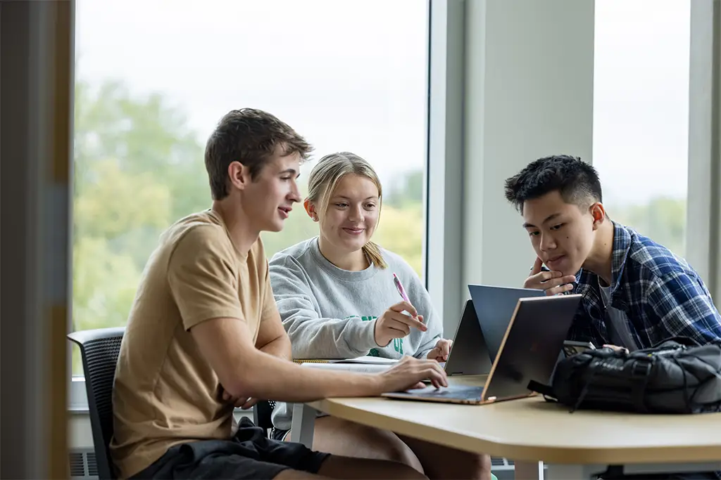 Three university students seated around a table in a bright classroom, collaborating on laptops and discussing whether to choose an ABET-accredited program for their studies