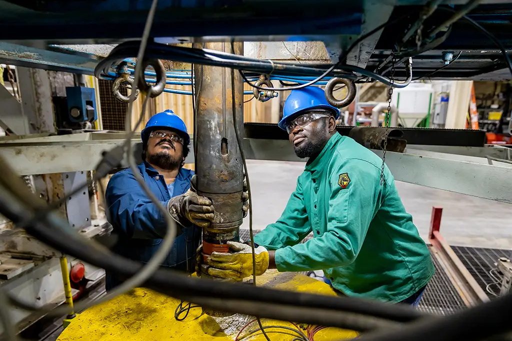 Two petroleum engineering students wearing hard hats and safety gear while performing hands-on work with industrial drilling equipment.