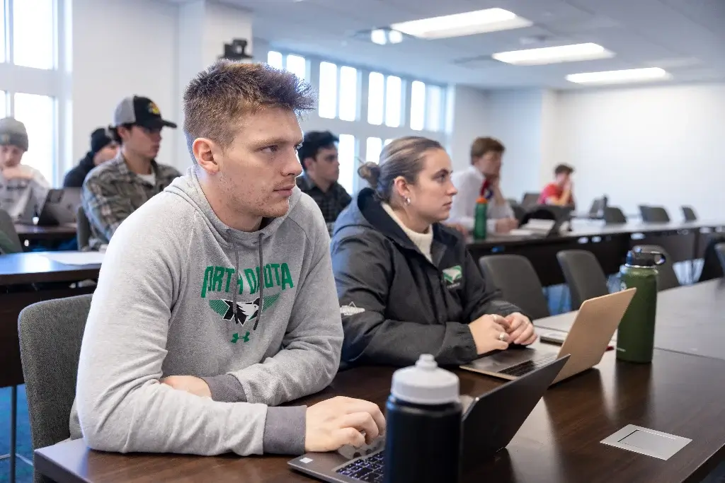 Classroom full of students engaged in business management lecture, using laptops.