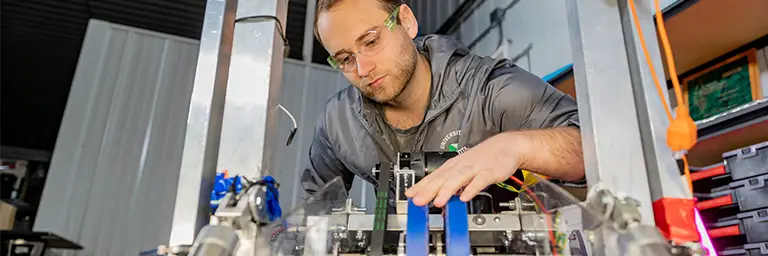 An engineering student wearing safety glasses works on the mechanical assembly of a complex robot in a university laboratory.