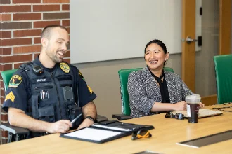 A DEA agent in uniform, holding a clipboard, and a civilian with a beverage container are engaged in a professional meeting or interview setting