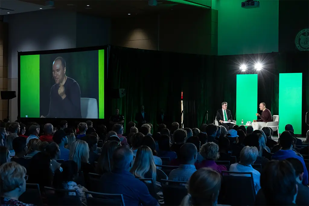 Large audience attending a conference session where two speakers discuss AI ethics on stage under green lighting, symbolizing the collaboration among governments, researchers, and tech leaders in shaping ethical AI.
