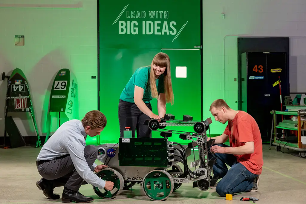 Three mechanical engineering students collaborating to assemble and test their four wheeled robotic rover design in the university lab.