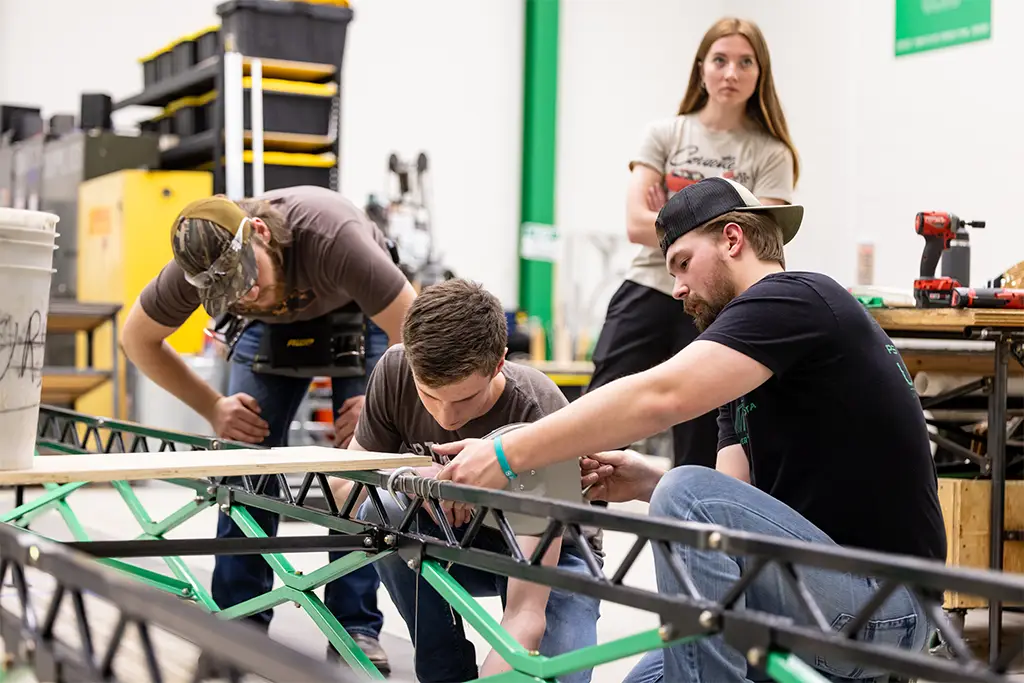 Engineering students work together in a university workshop to assemble a metal bridge structure. One student uses a drill while others assist with alignment and measurement, applying classroom concepts to a real-world project.