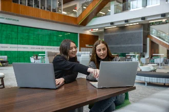 Two female business management students, with their laptops open, engaged in a discussion about their coursework