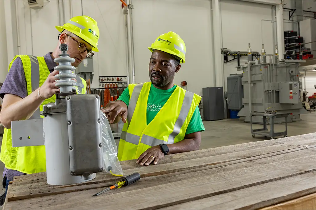 An experienced engineer mentors a student during an internship, illustrating the critical hands-on learning that complements a bachelor's degree. In a workshop, they collaborate over a piece of electrical equipment, applying classroom theory to a practical, real-world scenario.