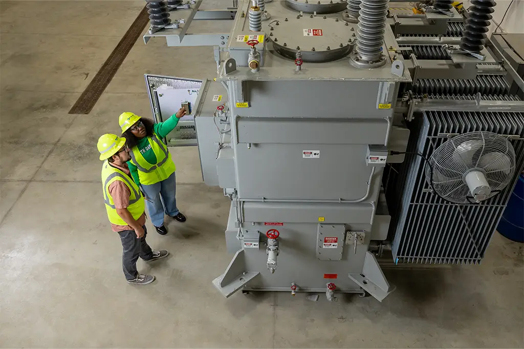 Two engineering students wearing safety vests inspect a large industrial machine inside a spacious facility. One student points at the machine while the other observes closely, demonstrating hands-on learning and teamwork.
