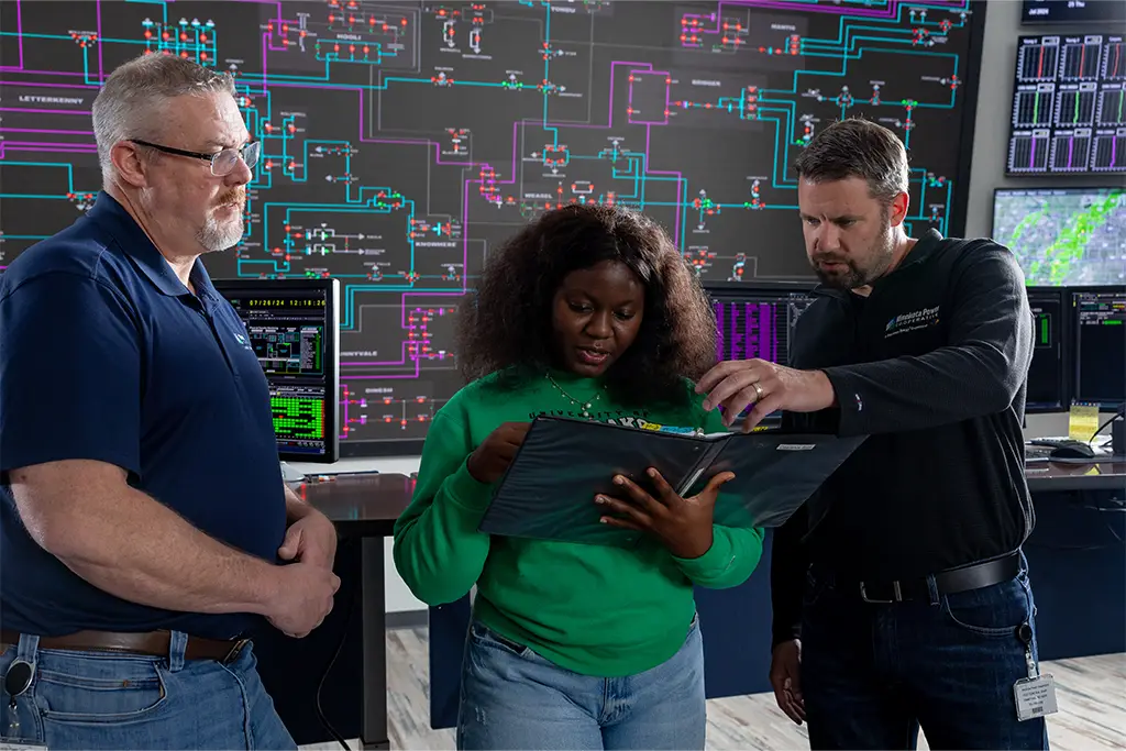 In a high-tech control room, two experienced engineers mentor a younger colleague, guiding her through technical plans in a binder. They stand in front of a large wall of complex system schematics, demonstrating hands-on collaboration and professional engineering work.